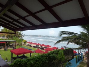 A beachside restaurant with outdoor seating and red umbrellas, overlooking the ocean.