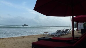 A beach with lounge chairs and a red umbrella, a sign for 'Sun Lesson', and a view of the ocean. - Samaru Beach House (Weligama)