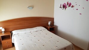 A bedroom with a wooden headboard, a bed with a patterned bedspread, a bedside table with a lamp, and a wall decoration of pink flowers. - Hostal Mary (Lloret de Mar)
