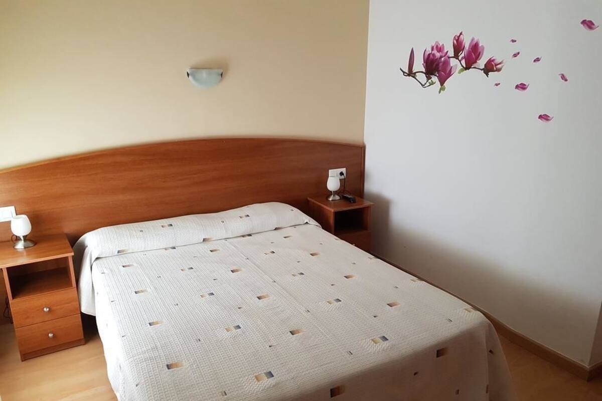 A bedroom with a wooden headboard, a bed with a patterned bedspread, a bedside table with a lamp, and a wall decoration of pink flowers.