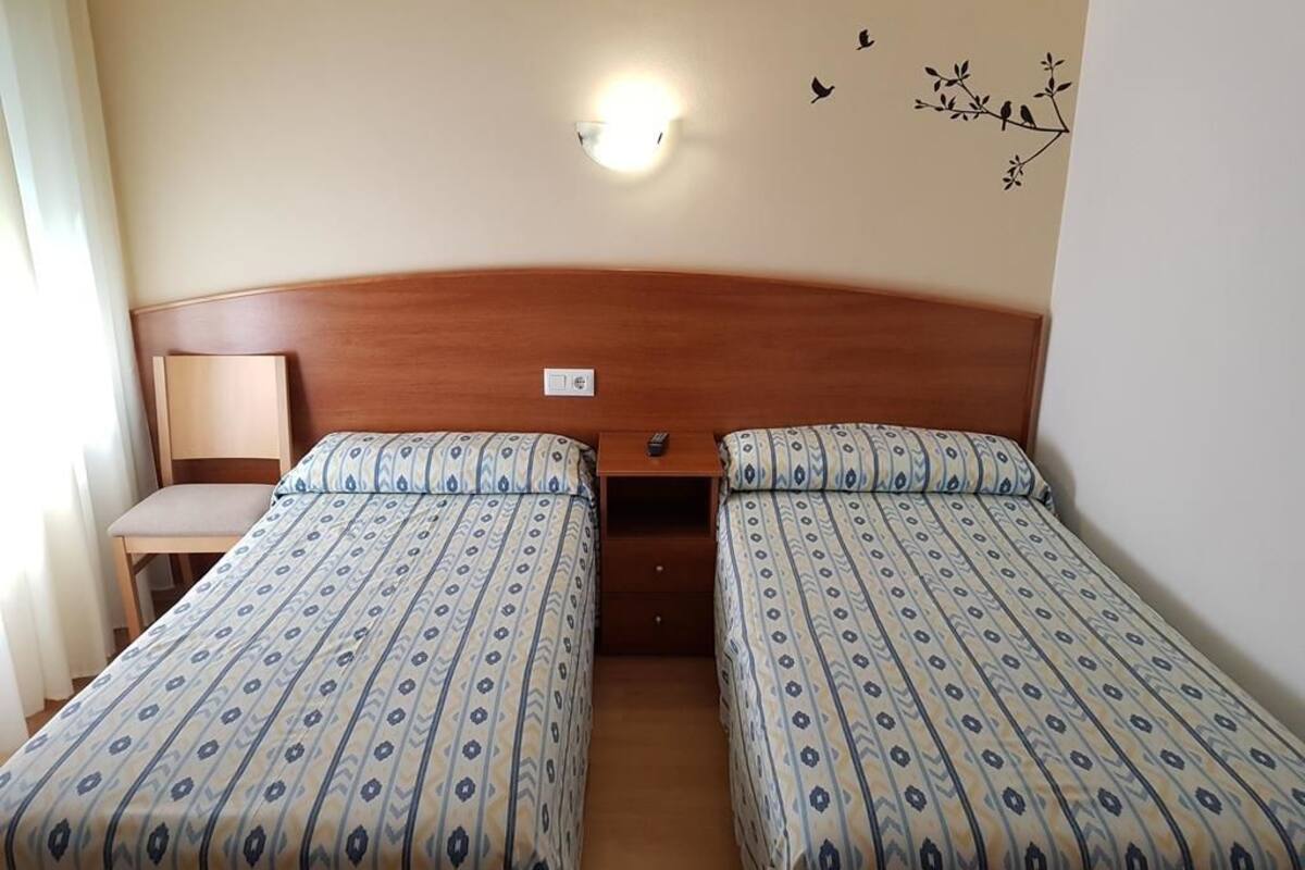 A hotel room with two single beds, a wooden headboard, a bedside table with a lamp, and a wall-mounted light fixture.