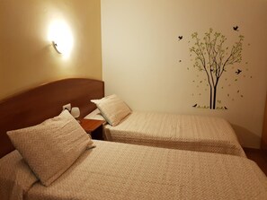 A hotel room with two beds, each with a patterned bedspread and pillows. There is a wall-mounted light fixture on the left and a wall decal of a tree with birds. - Hostal Mary (Lloret de Mar)