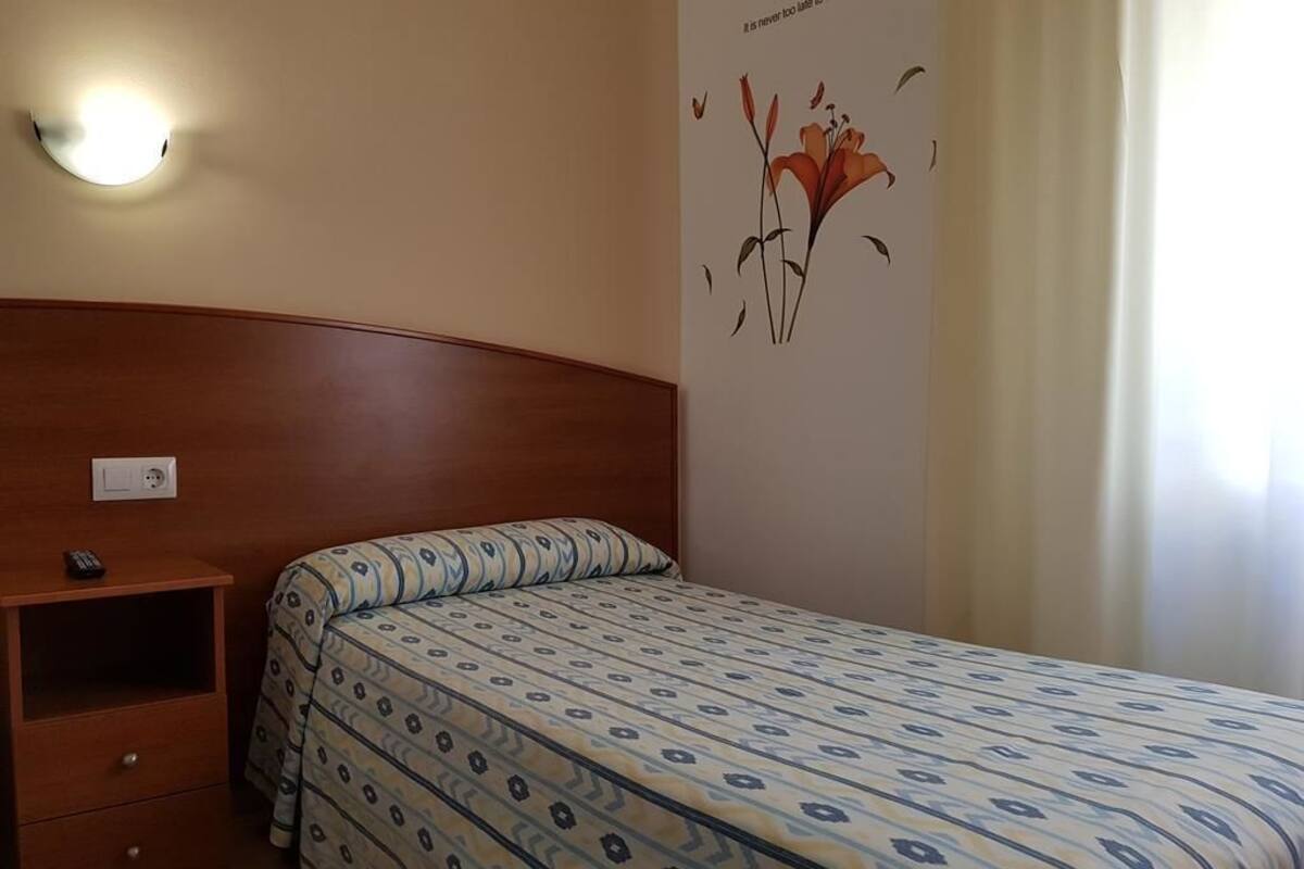 A hotel room with a wooden headboard, a bed with patterned bedding, a bedside table with a remote control, and a wall-mounted light fixture.