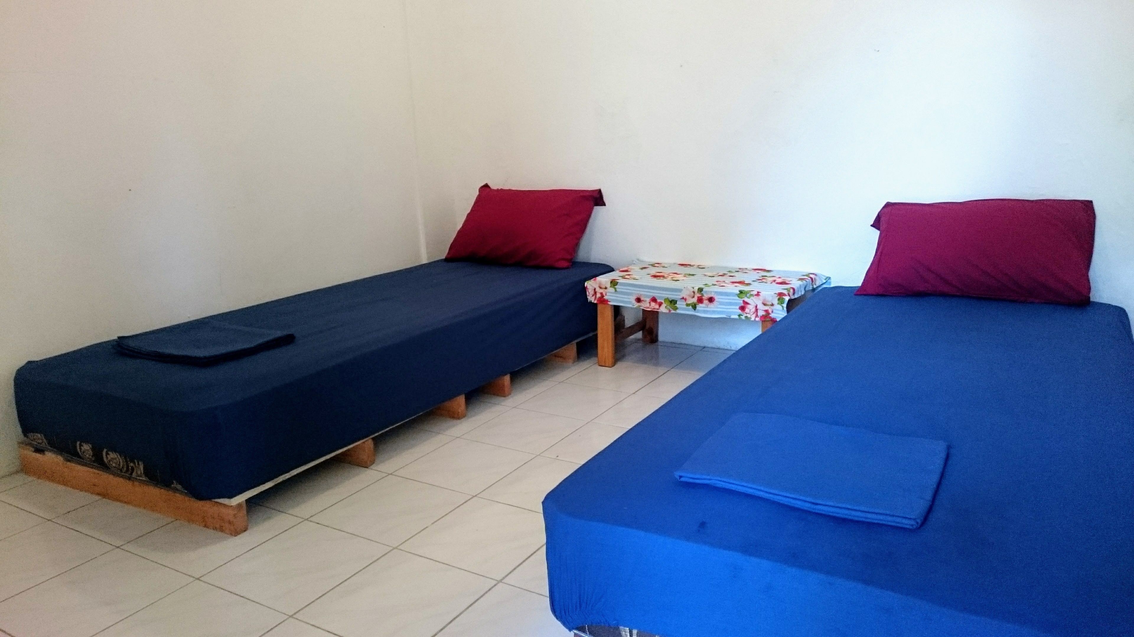 A room with two single beds, each with a blue mattress and a red pillow. There is a small table with a floral tablecloth between the beds.