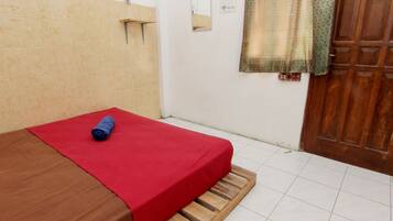 A small, simple room with a bed, a wooden bench, a red bedspread, a mirror, and a door.