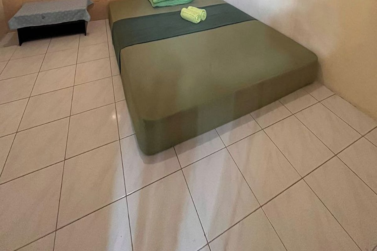 Una habitación pequeña y limpia con una cama individual, un banco y un ventilador de pared.