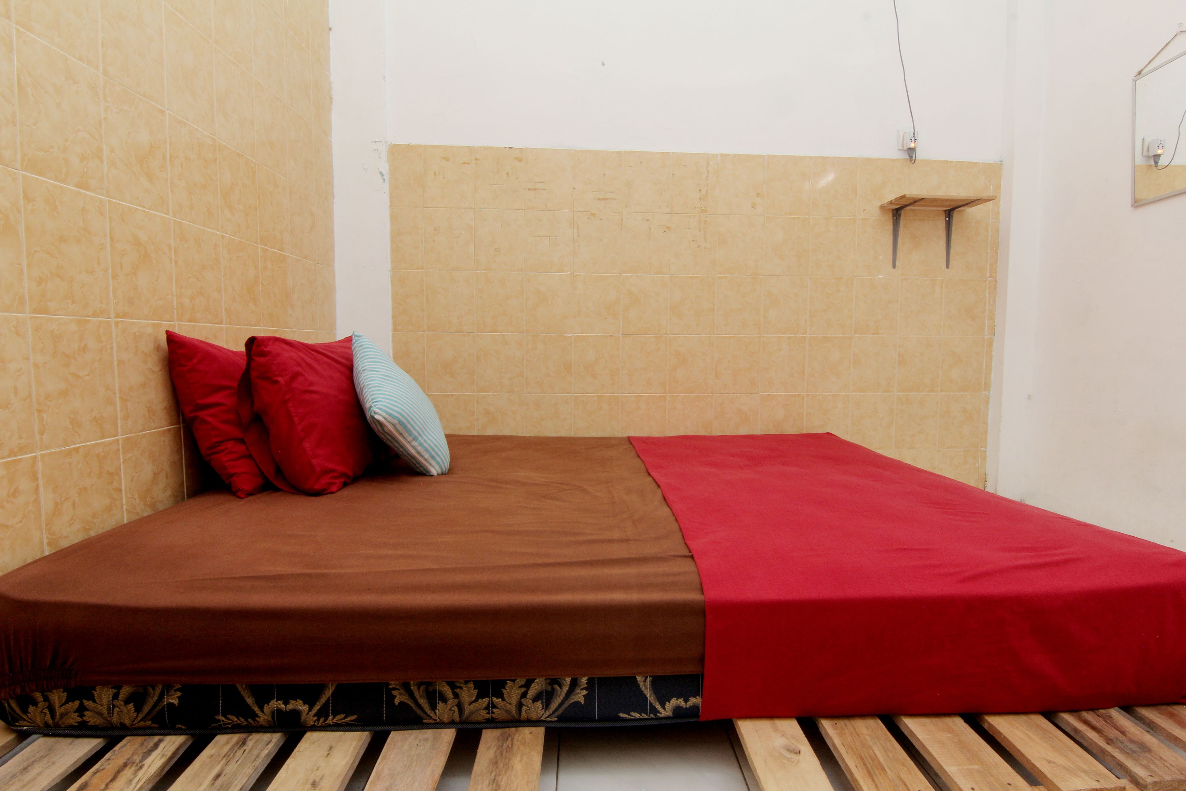 Een eenpersoonsbed met een bruine matras en rode en blauwe kussens, een houten hoofdbord en een rode deken.