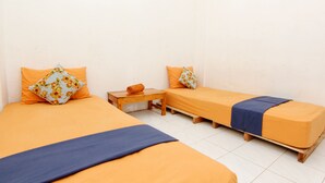 Ein Zimmer mit zwei Betten, jeweils mit einer orangefarbenen Tagesdecke und einer blauen Decke. Zwischen den Betten steht ein Holznachttisch mit einem zusammengerollten Handtuch und ein kleiner Tisch.