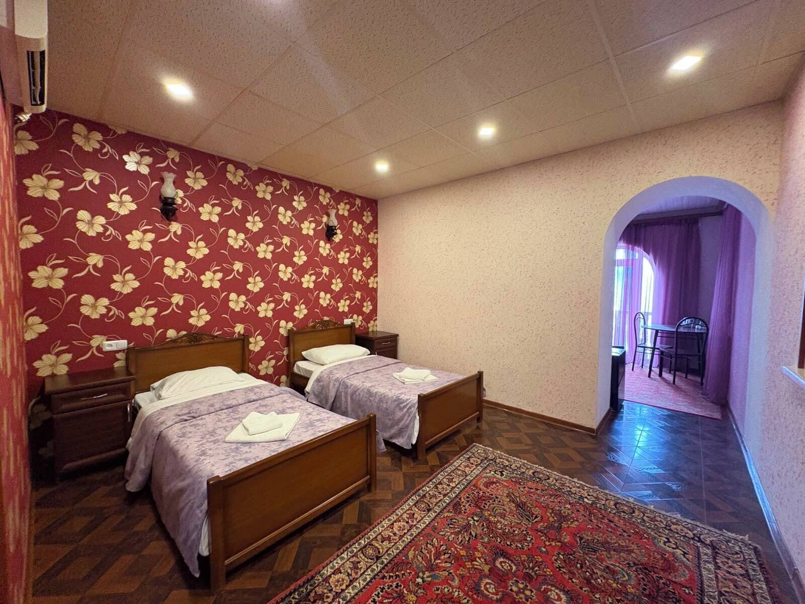 Quarto com duas camas de madeira, um tapete estampado, papel de parede floral e um corredor que dá para uma sala de jantar.