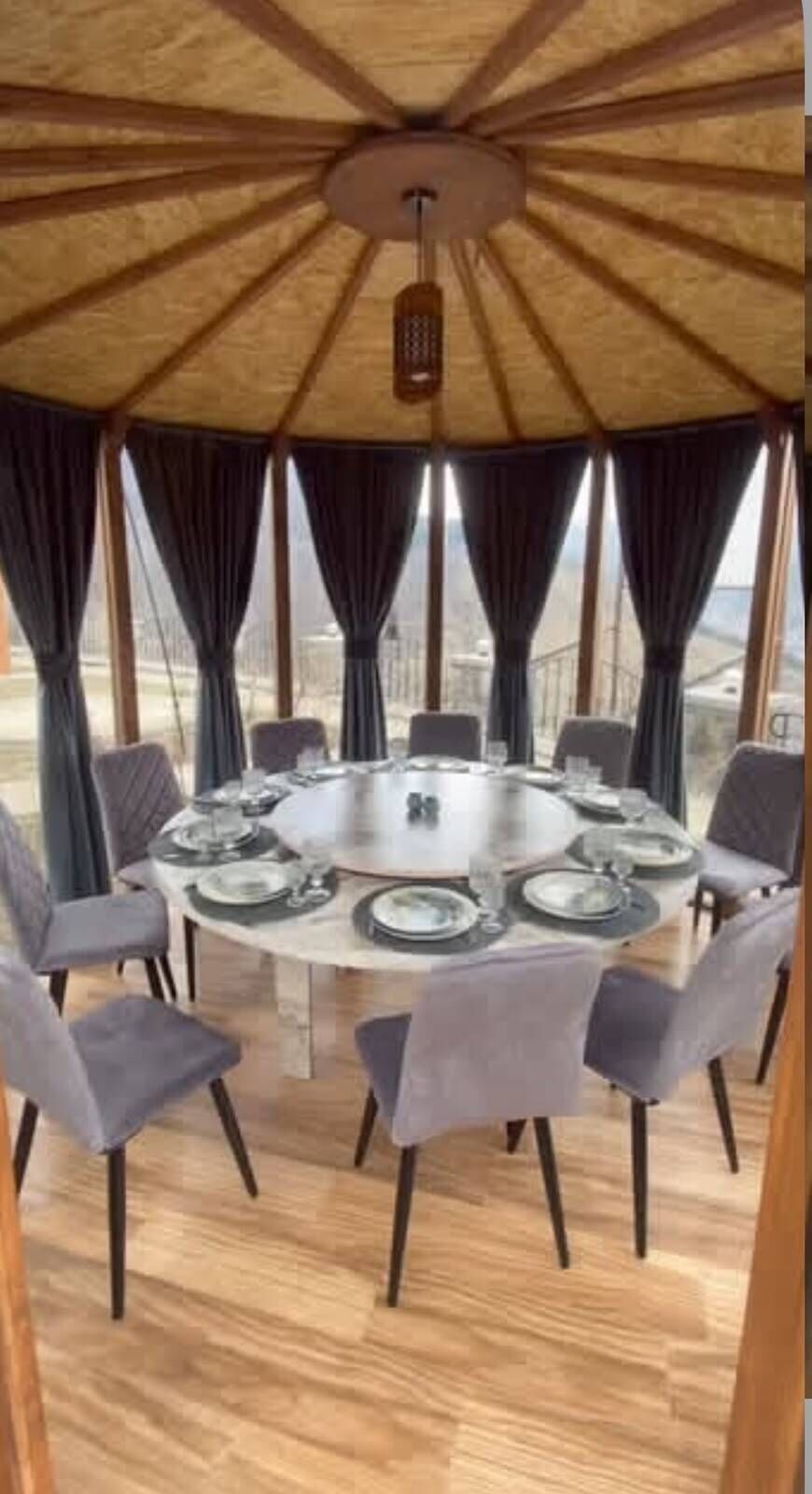 Una mesa redonda con sillas, preparada para una comida, bajo un techo de madera con una lámpara colgante.