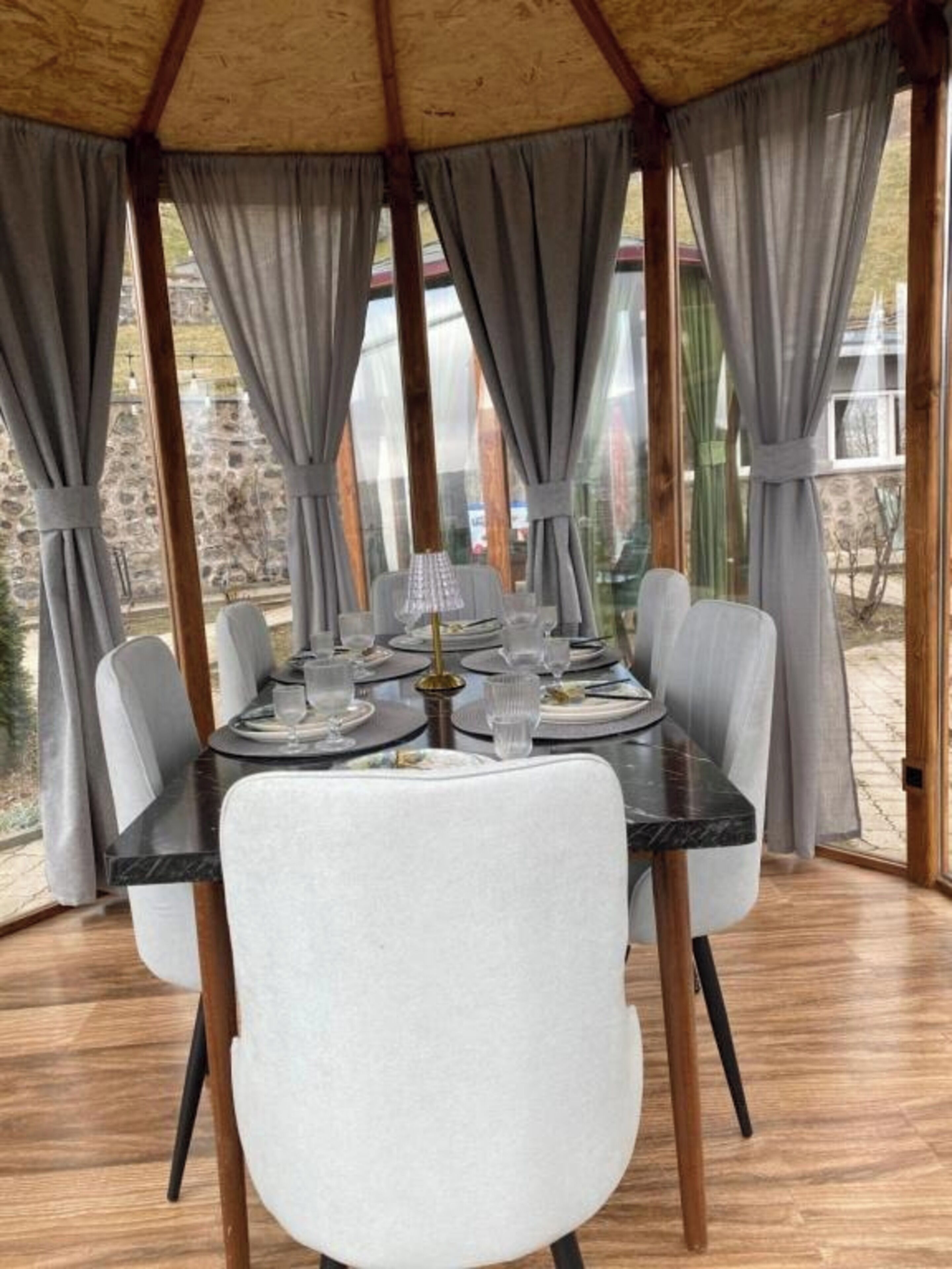 Un coin repas avec une table dressée pour quatre personnes, entourée de chaises, sous une structure de plafond en bois agrémentée de rideaux.