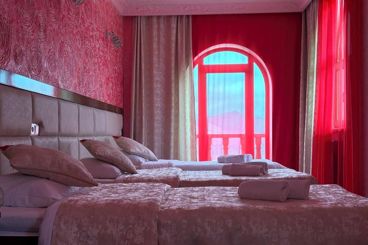 Una habitación de hotel con una cama grande, una cama más pequeña y un sofá cama. Se ve una ventana con cortinas rojas y vistas a un edificio.