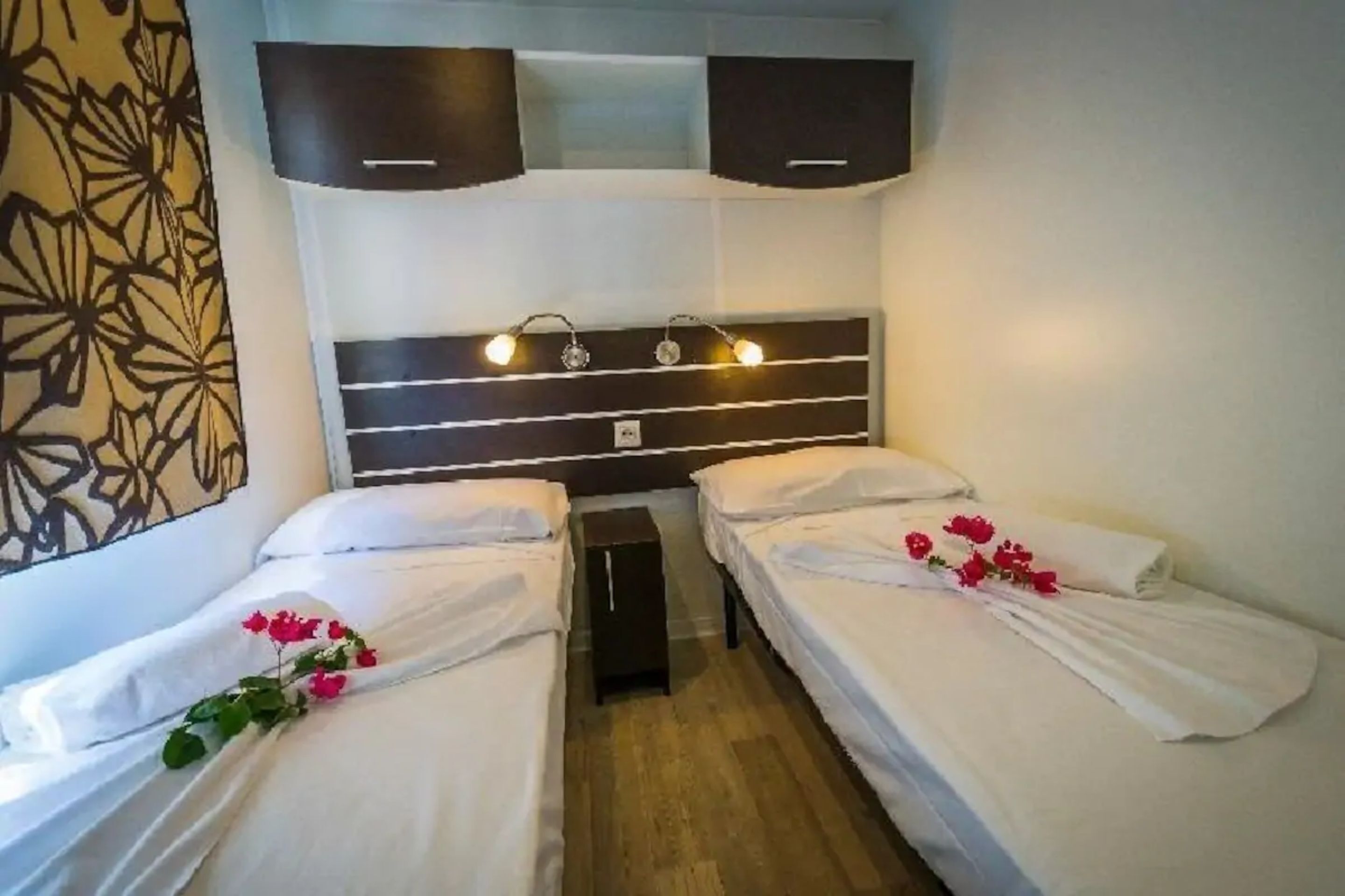 Quarto de hotel com duas camas de solteiro, piso de madeira, cabeceira moderna com iluminação embutida e um objeto decorativo na parede.