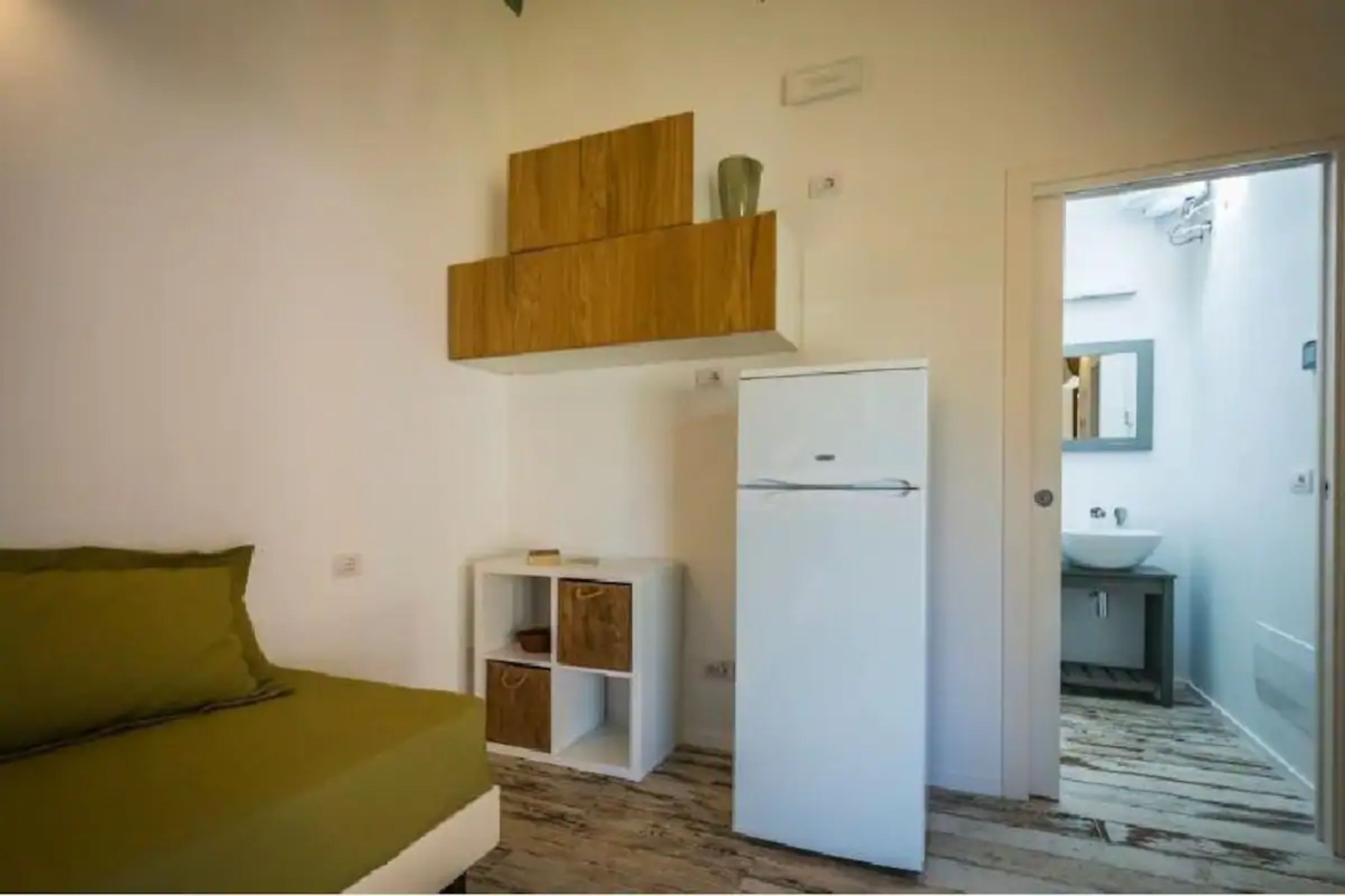 A compact room with a bed, a white refrigerator, a small wooden cabinet, and a door leading to a bathroom.