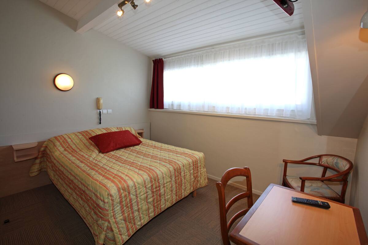 Un dormitorio con cama, mesa y silla. Hay una ventana con cortinas y una lámpara en la pared.