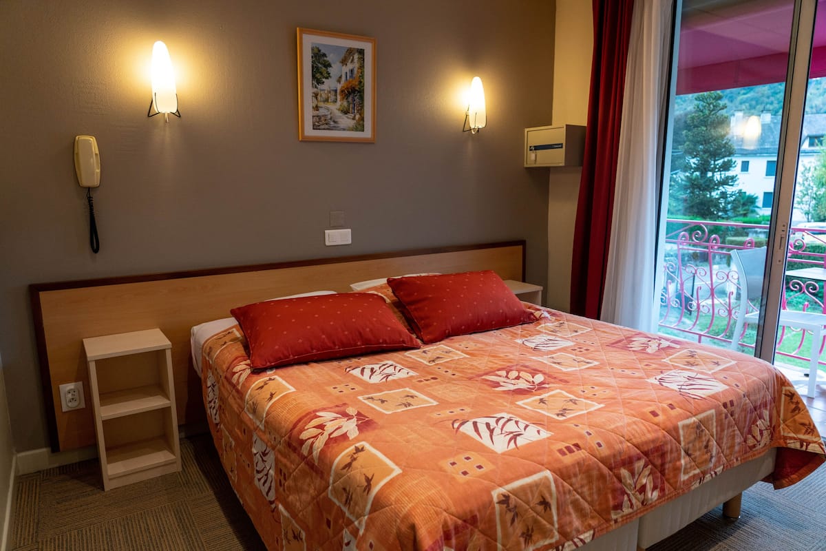 Habitación de hotel con cama, dos lámparas de noche, un teléfono, un estante pequeño y ventana con vistas al balcón.