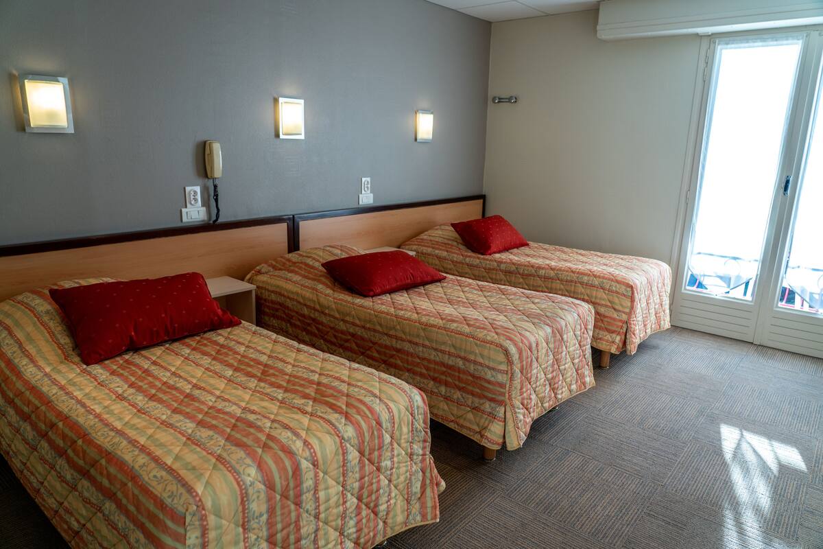 Habitación de hotel con tres camas, cada una con almohada roja y edredón estampado. Hay luces fijadas en la pared sobre cada cama y un teléfono en la pared.