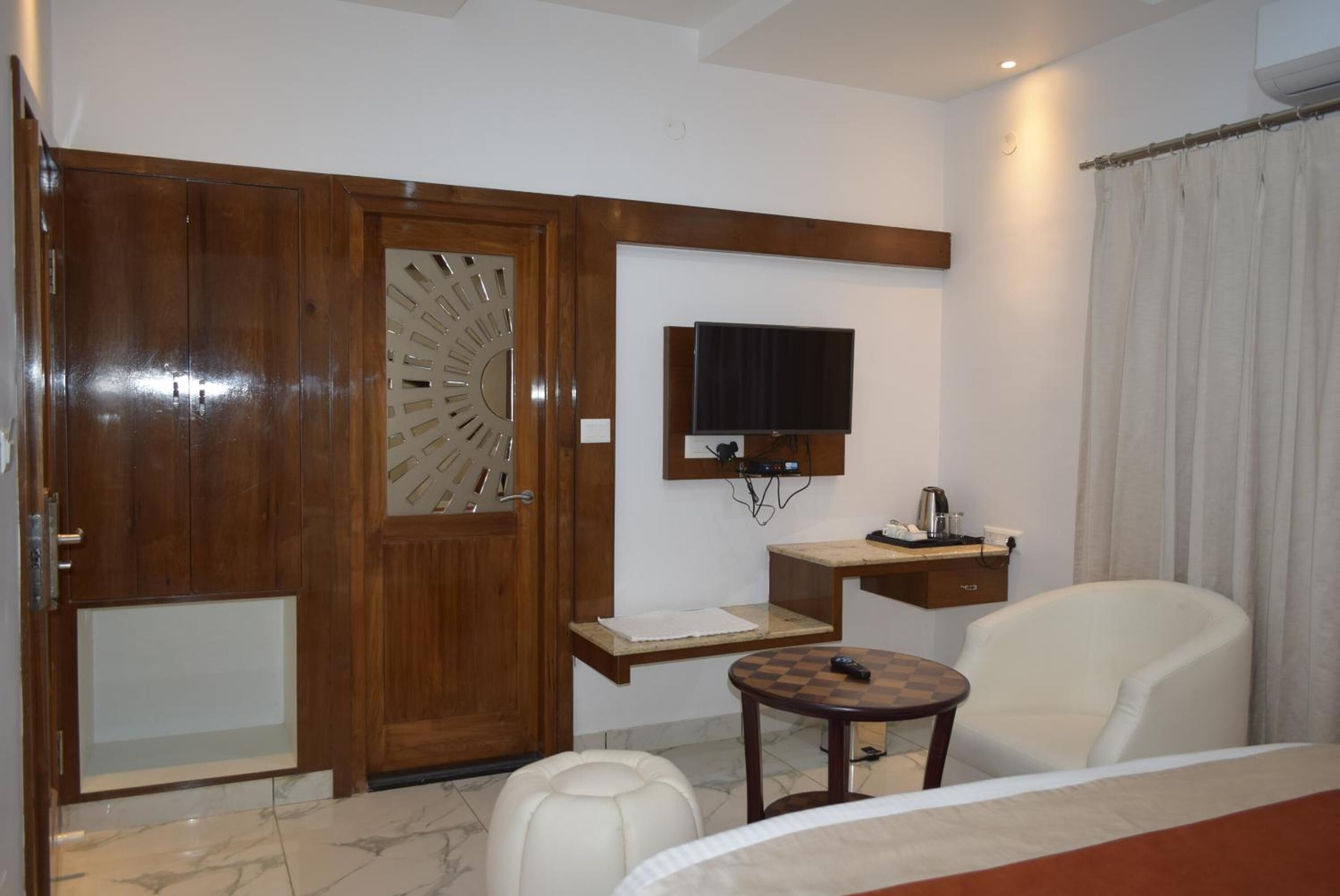 A hotel room with a wooden wardrobe, a television mounted on the wall, a small table with a remote control, a white armchair, and a bed.