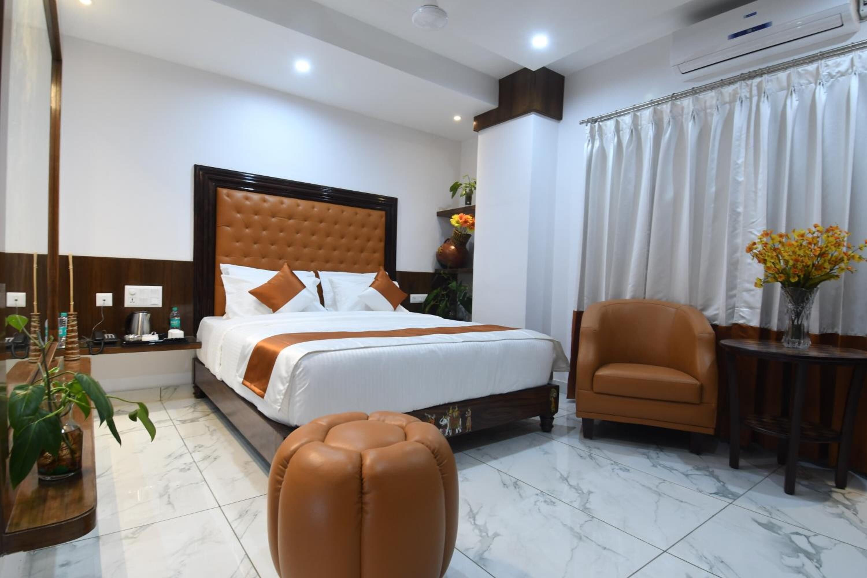 A hotel room with a large bed, a brown leather armchair, a small table with a vase of flowers, and a mirror.