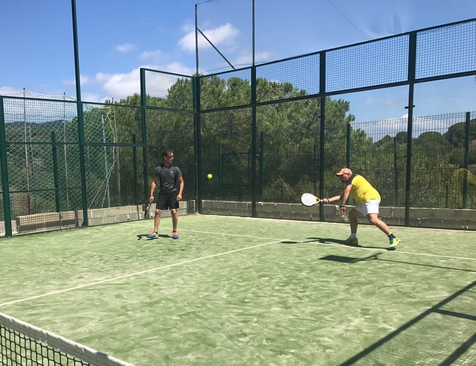 An outdoor tennis court with a net, surrounded by a green fence and trees.