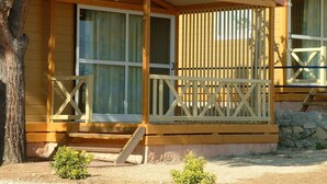 A wooden cabin with a porch, large windows, and a stone foundation. - Camping Roca Grossa (Calella)