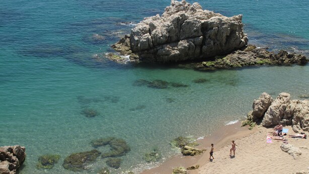 A beach with clear water, rocky formations, and people sunbathing. - Camping Roca Grossa (Calella)