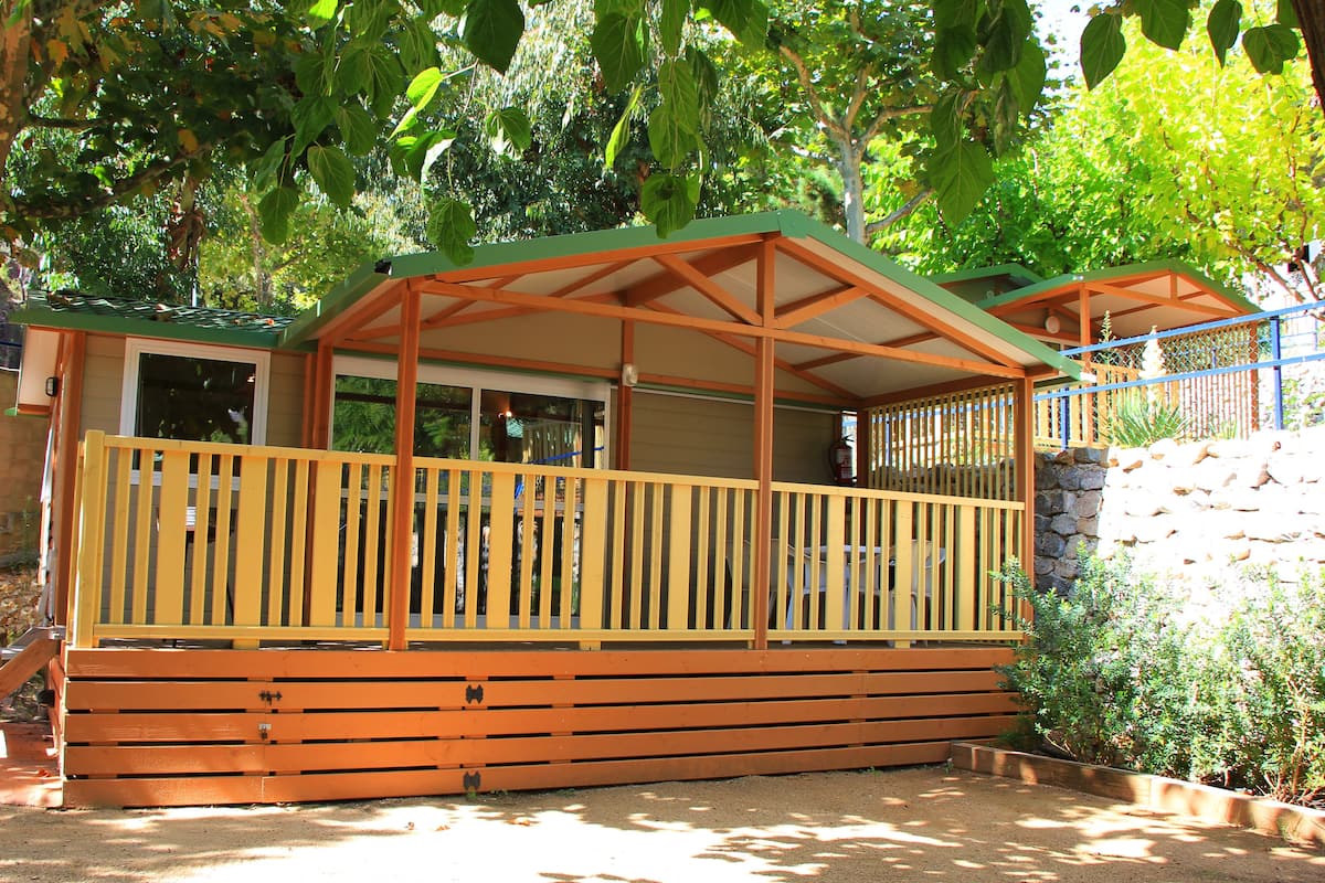 A wooden cabin with a balcony, surrounded by trees and a stone wall.