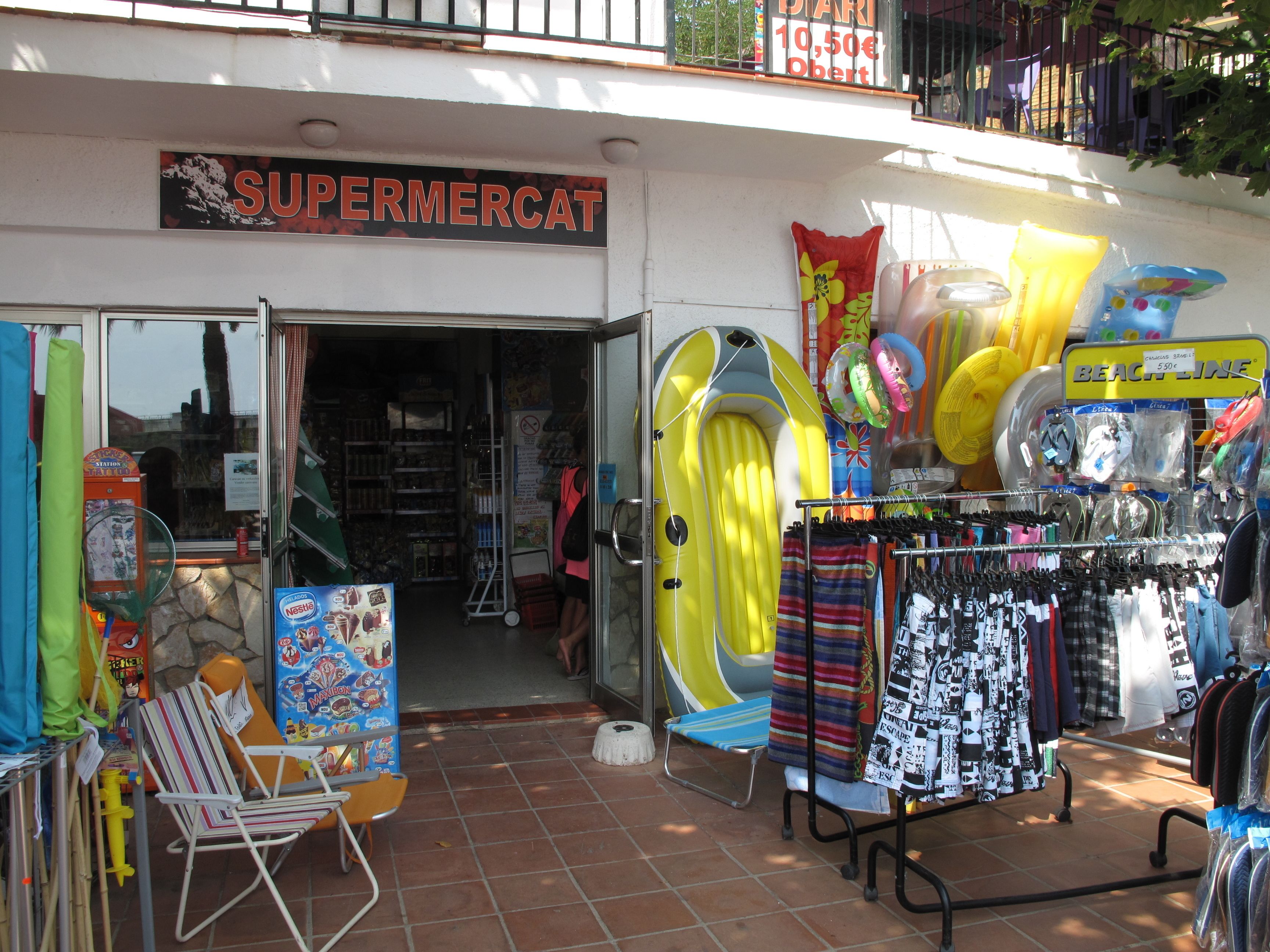 A storefront with a sign reading "SUPERMERCAT" and various outdoor items like inflatable rings and beach toys displayed outside.