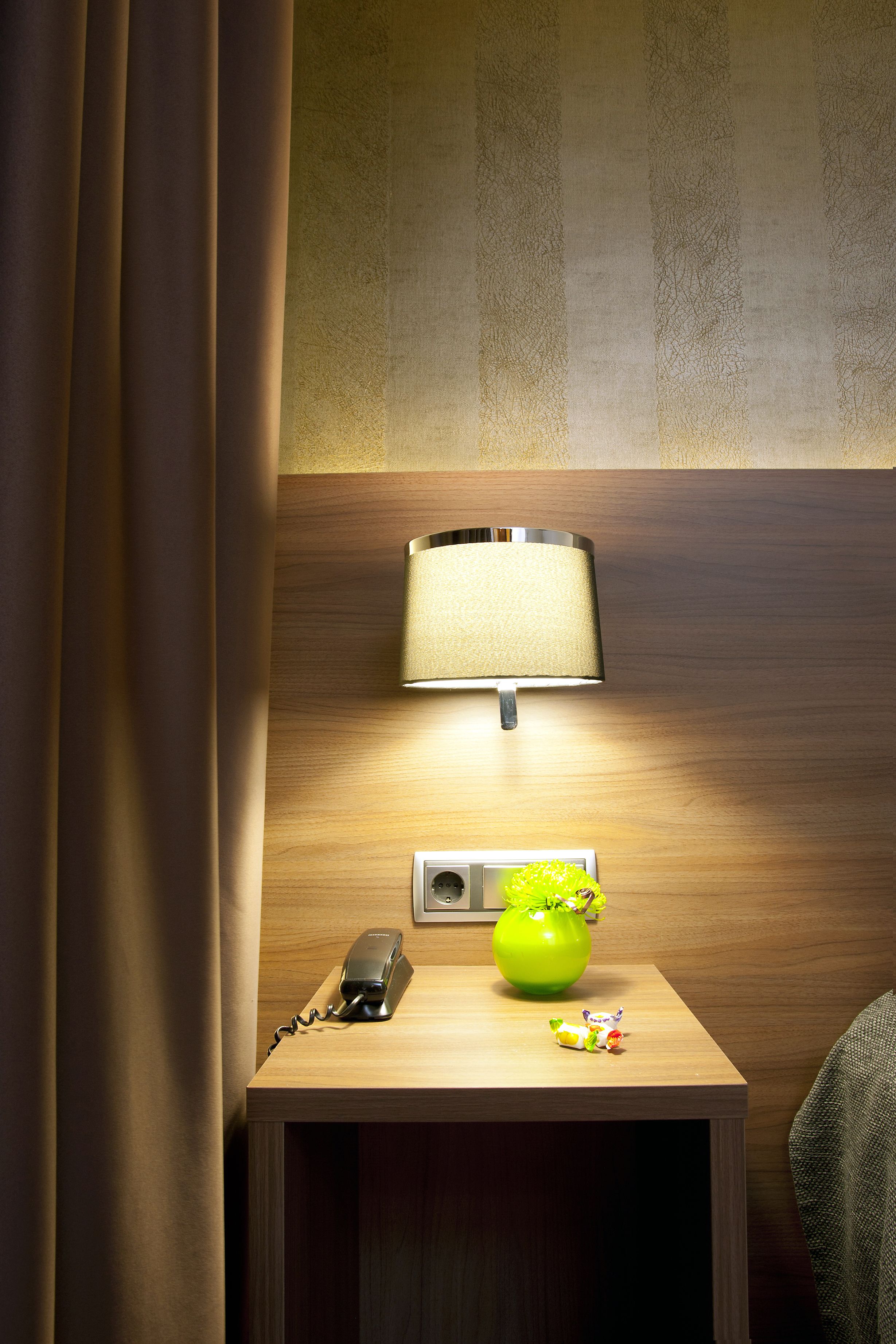 A wooden bedside table with a lamp, a telephone, and a green apple vase.