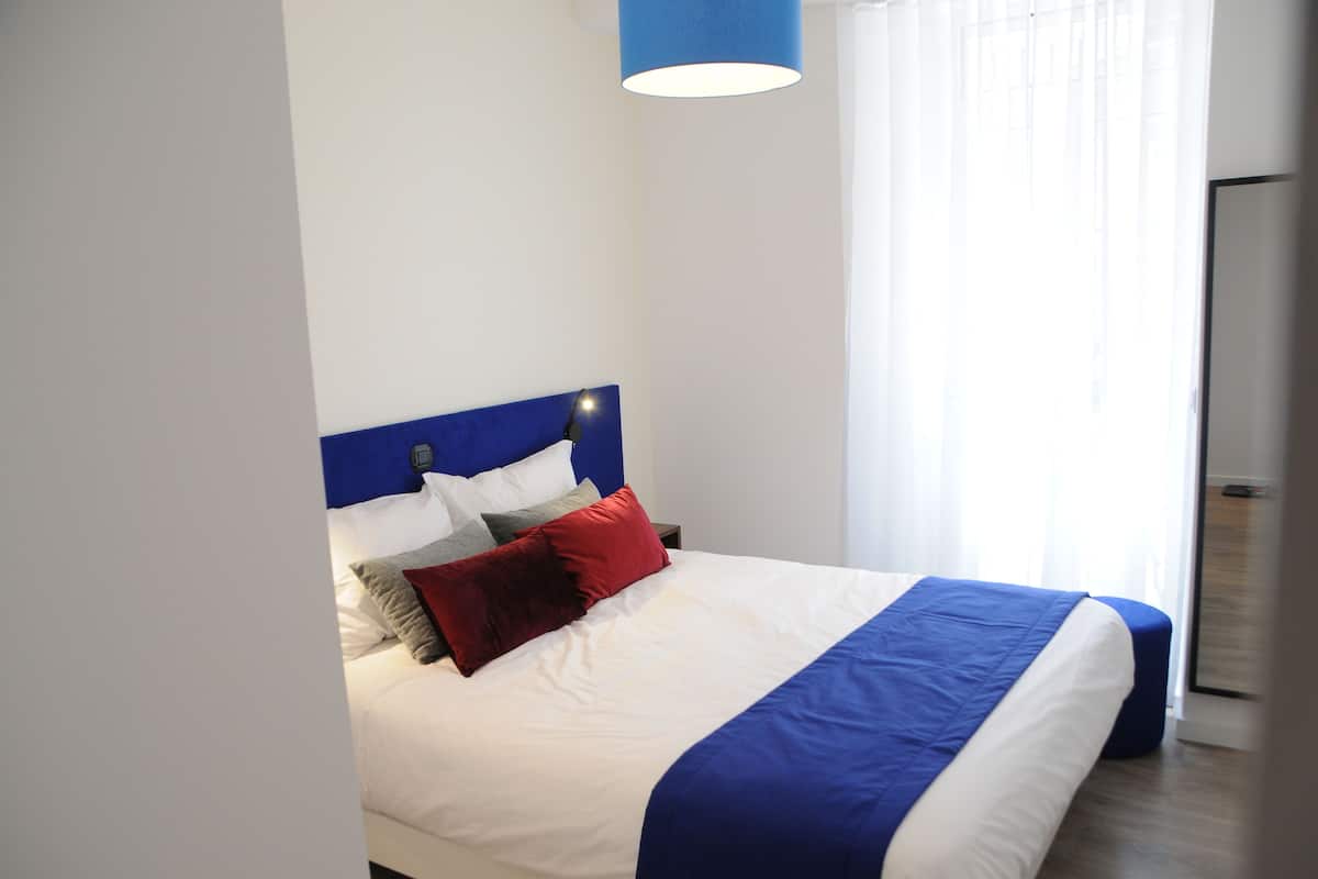 Un lit bien fait, avec une tête de lit bleue, du linge de lit blanc et un coussin rouge.