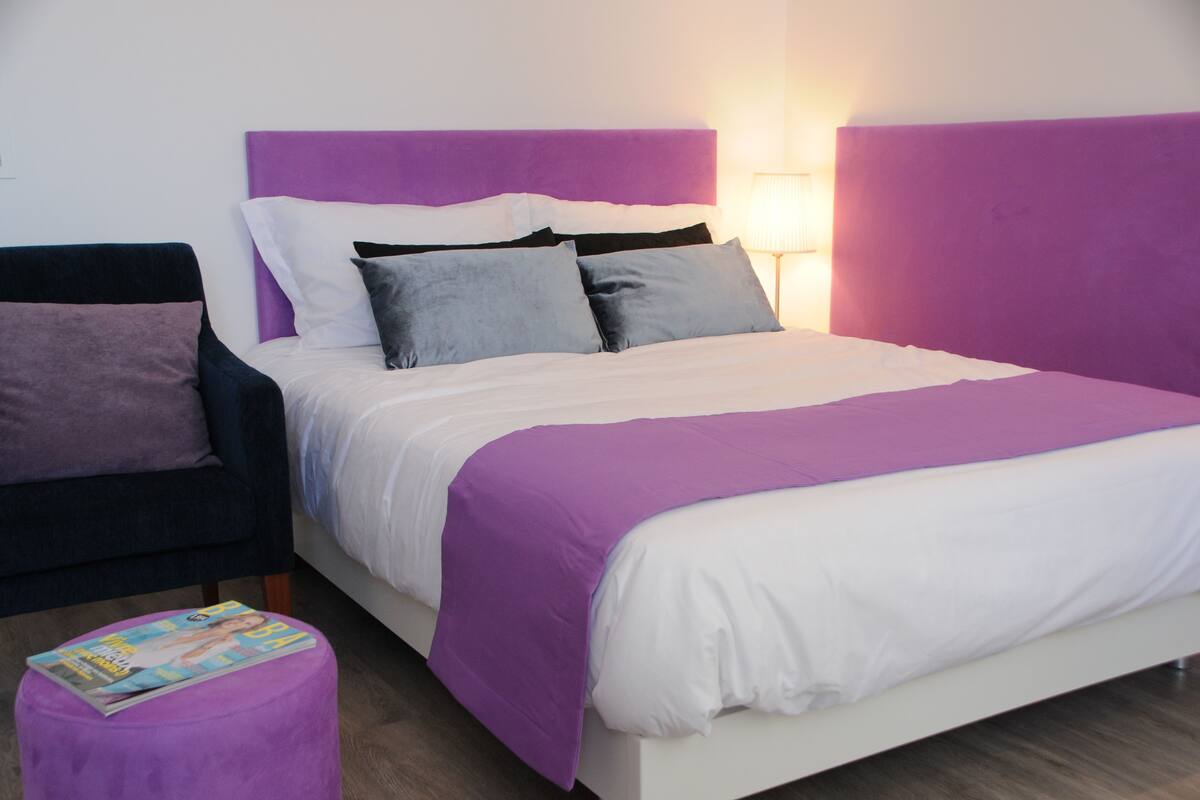Un lit avec une tête de lit violette, du linge de lit blanc et un magazine posé sur un pouf violet.