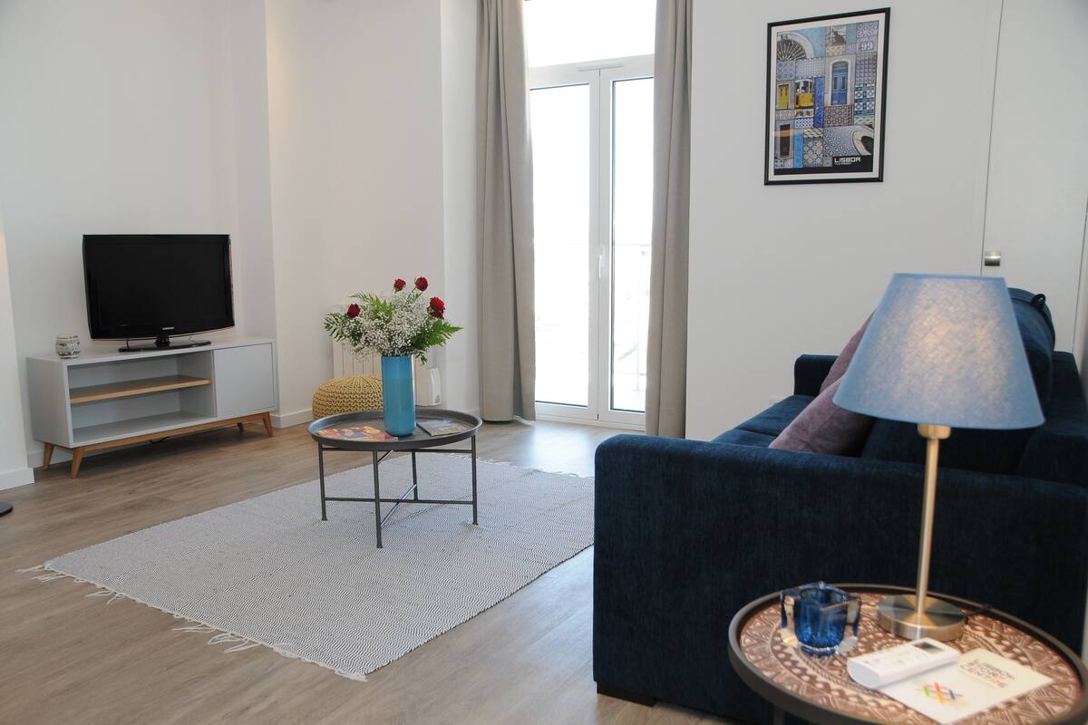 Un salon moderne avec un téléviseur à écran plat, un canapé bleu, une petite table d’appoint avec une lampe, un vase de fleurs et une grande fenêtre avec des rideaux.