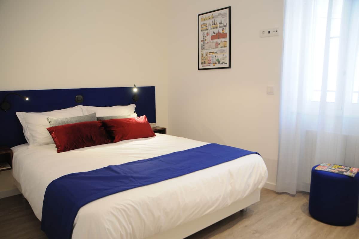 Un lit bien fait, recouvert d’une couvre-lit bleue et blanche, avec un oreiller rouge et une couverture bleue. On y trouve un petit pouf bleu sur lequel sont posés des magazines, et un tableau encadré accroché au mur.