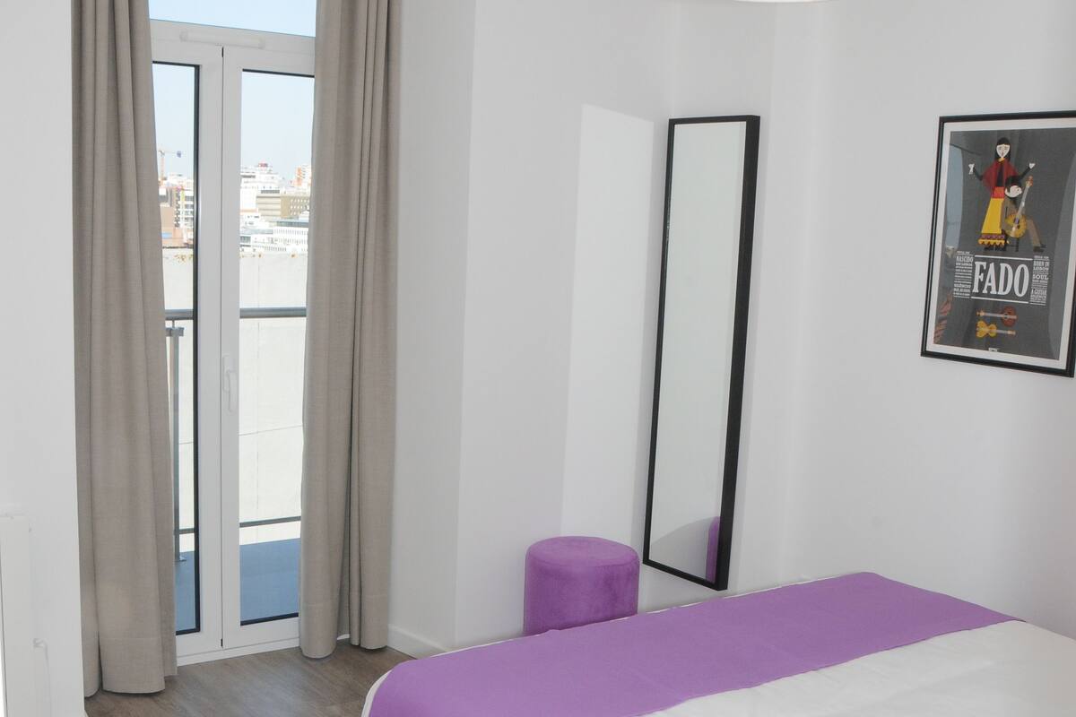 Une chambre à coucher avec un lit, une grande fenêtre avec des rideaux, un luminaire au plafond et un tableau encadré accroché au mur.