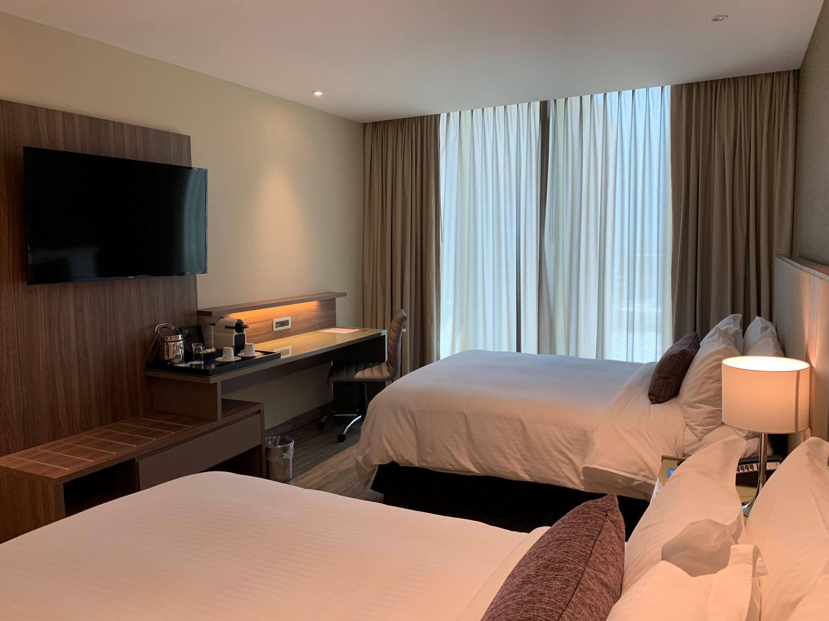 Habitación de hotel con dos camas, un escritorio con silla, un televisor montado en la pared y una ventana con cortinas.