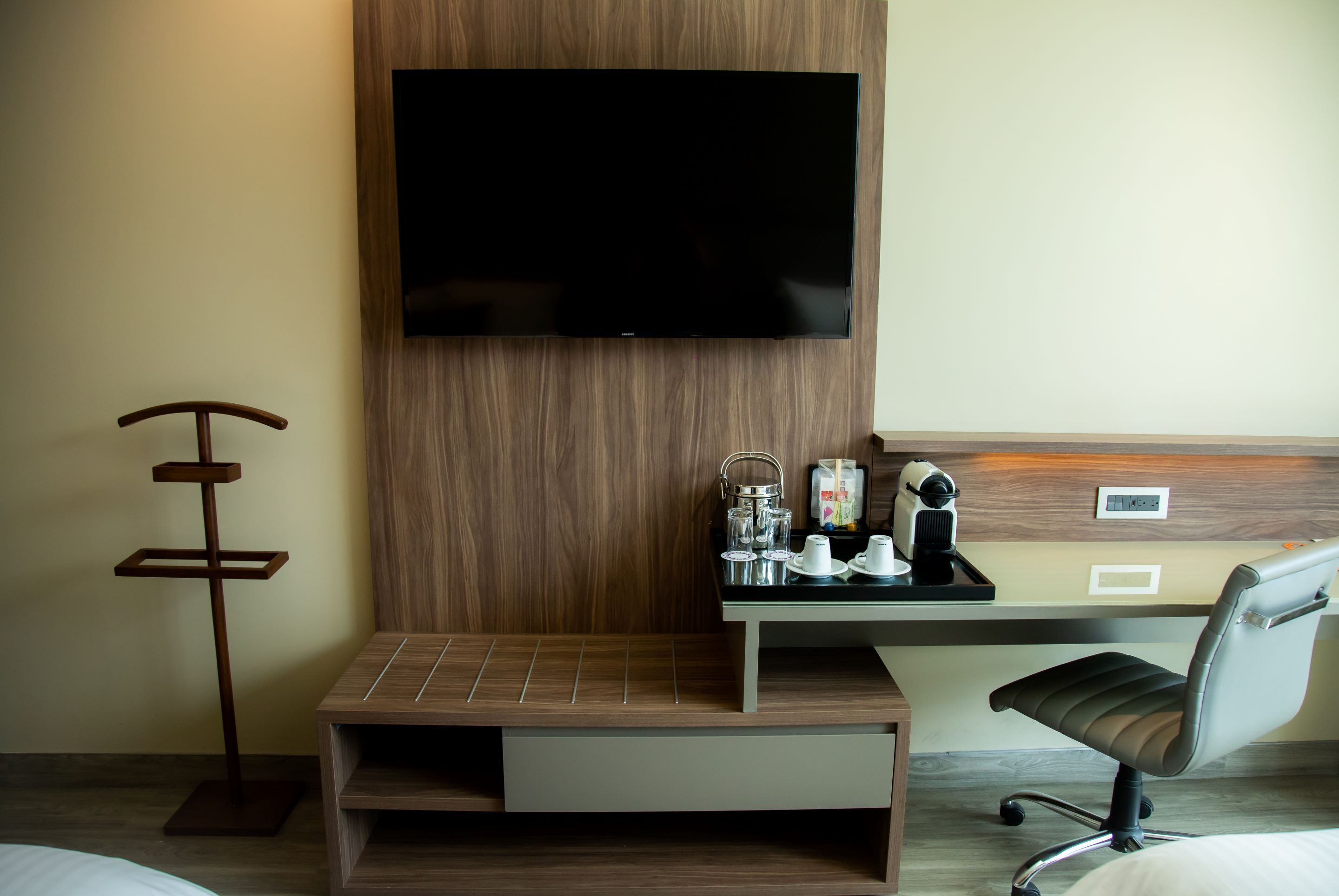 A hotel room with a flat-screen TV mounted on a wooden wall, a small table with a tray of breakfast items, a chair, and a bedside table.