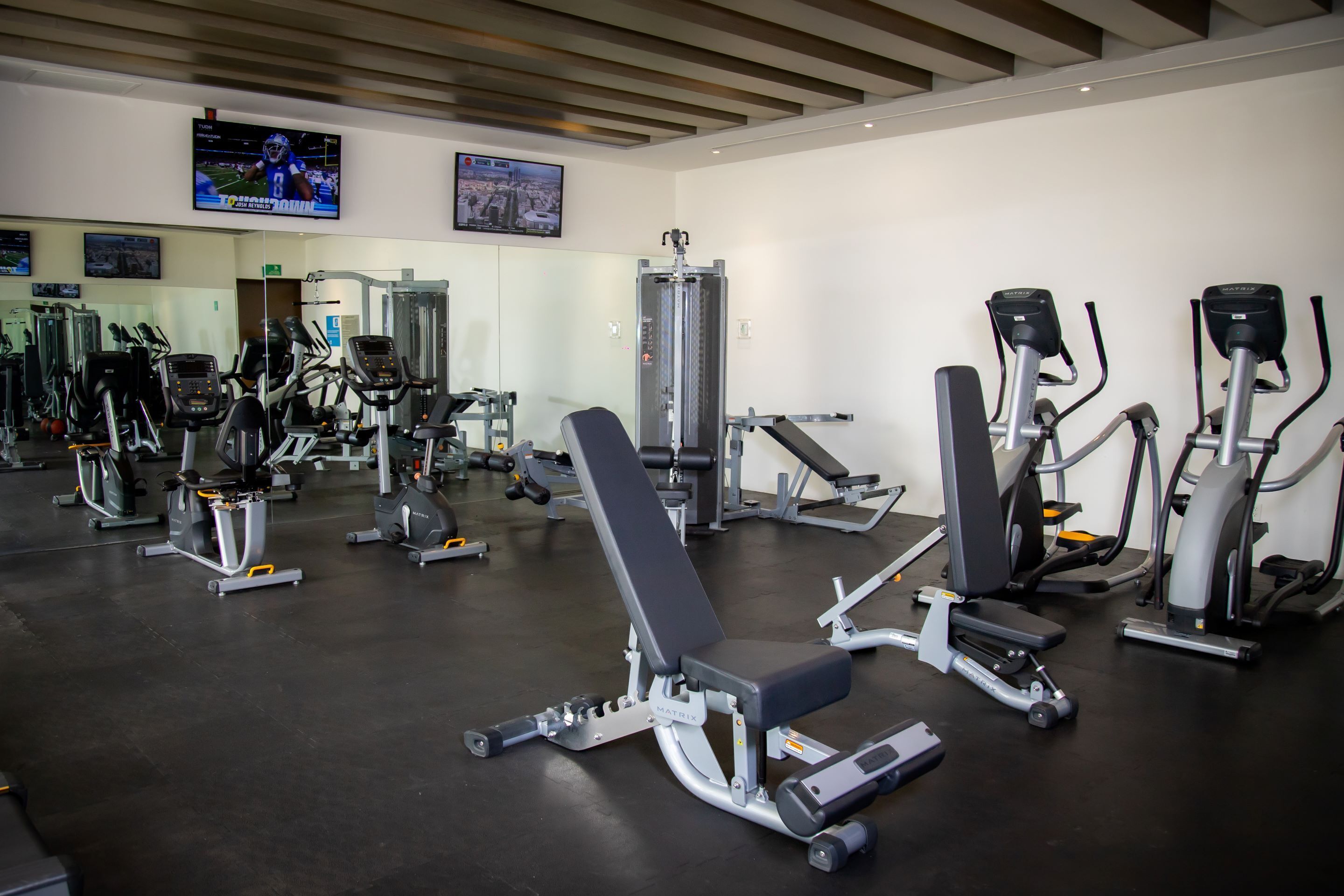 A gym with various exercise machines, a mirrored wall, and a television displaying a sports event.