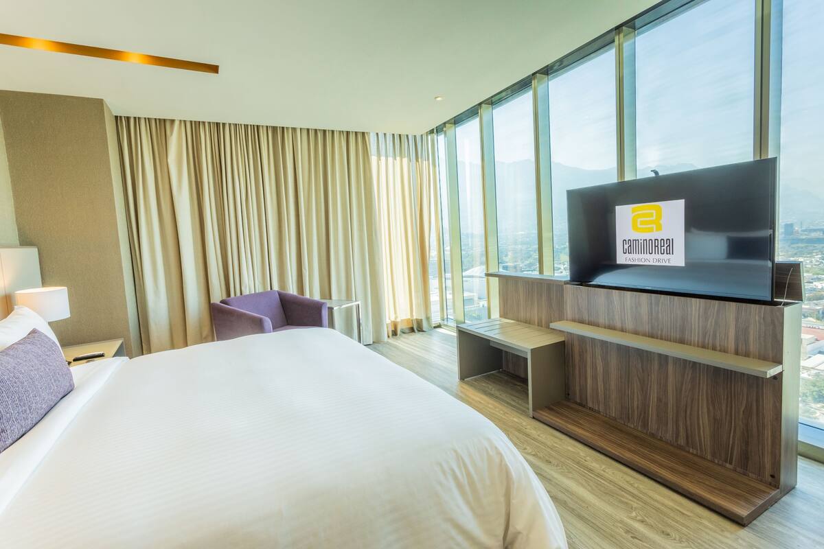 Une chambre d’hôtel dotée d’un grand lit, d’un téléviseur fixé au mur en bois, d’un bureau et offrant une vue sur la ville grâce à de grandes fenêtres.