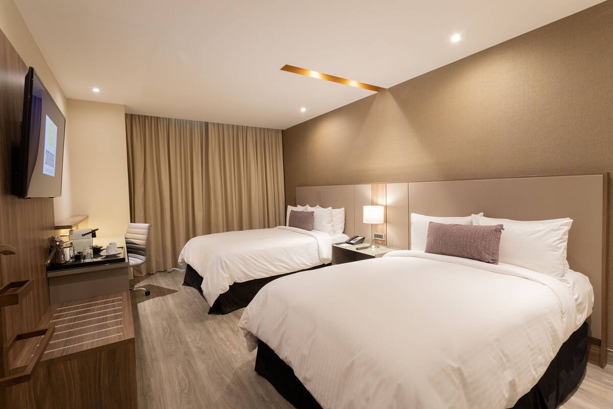 Ein Hotelzimmer mit zwei Betten, einem Schreibtisch mit Stuhl, einem Flachbildfernseher und einem großen Fenster mit Vorhängen.