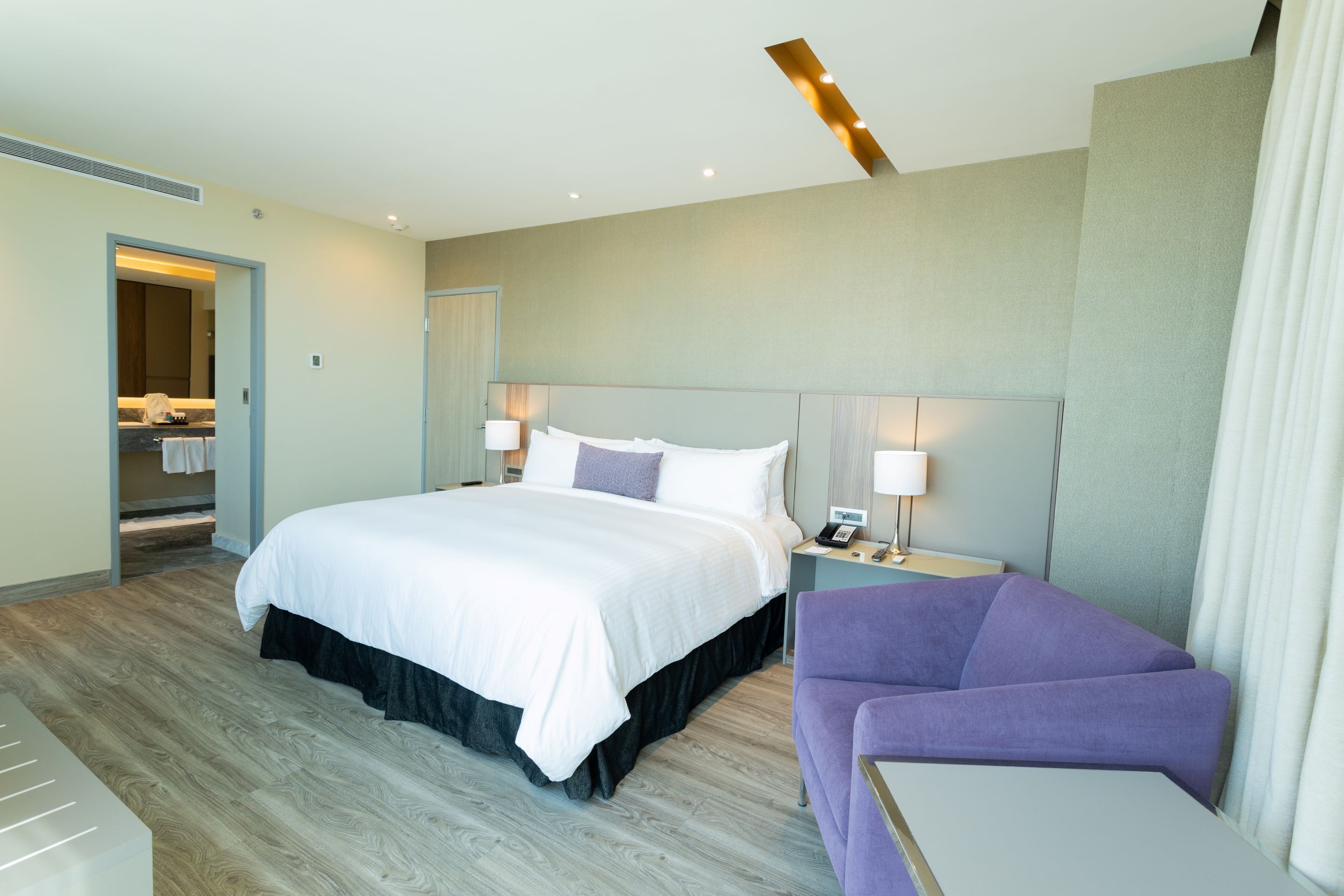 Une chambre d’hôtel moderne avec un grand lit, une table de chevet avec une lampe, un fauteuil violet et une vue sur la salle de bain par une porte ouverte.