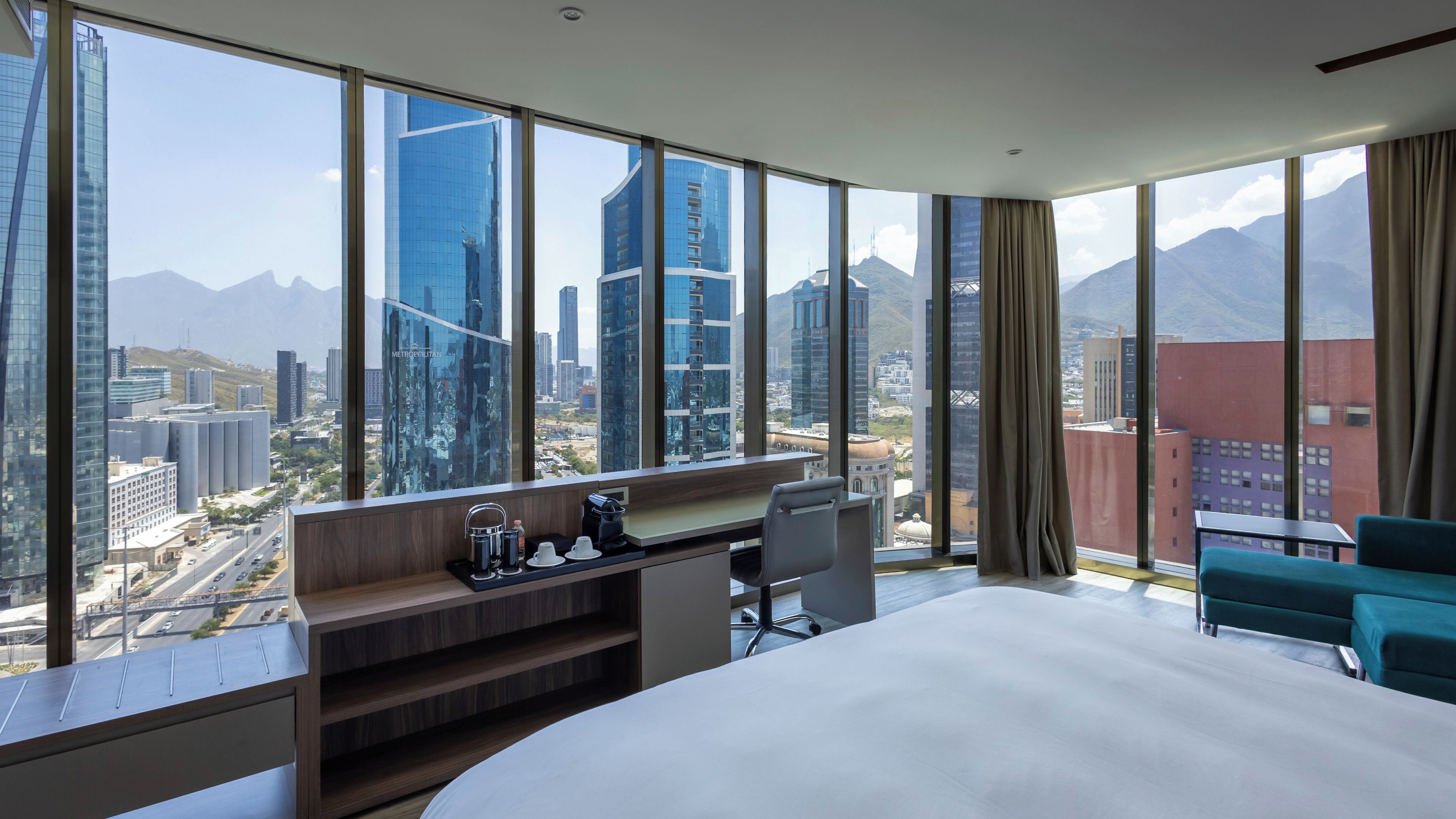A hotel room with a large bed, a desk with a chair, a coffee maker, and a view of a cityscape with tall buildings and mountains.
