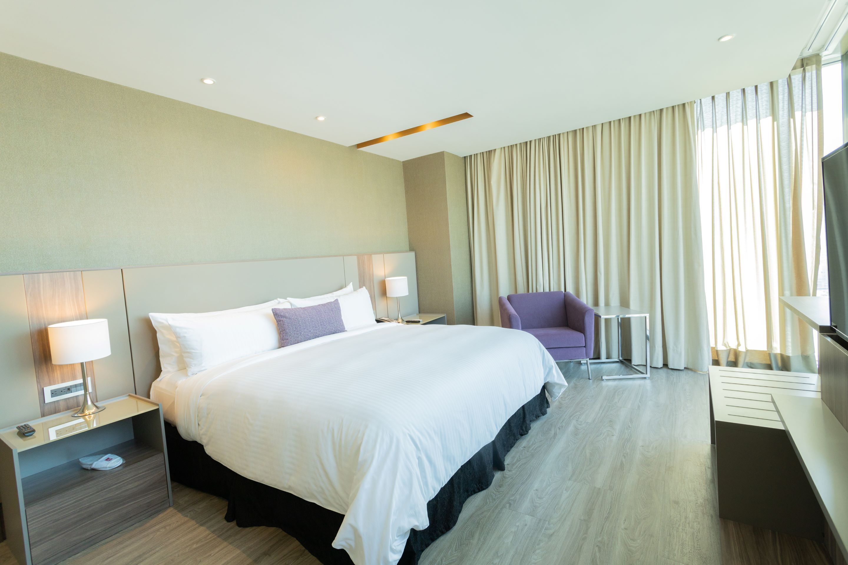 Une chambre d’hôtel moderne dotée d’un grand lit, de tables de chevet avec des lampes, d’un téléviseur à écran plat, d’un petit coin salon et d’une grande fenêtre avec des rideaux.