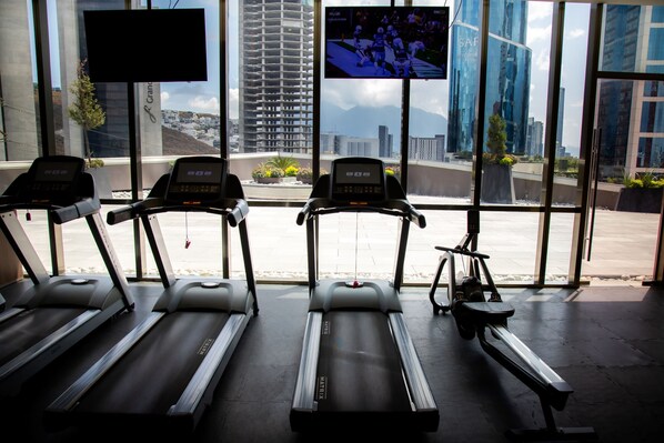A gym with treadmills and a stationary bike, large windows with a city view, and a TV screen displaying a sports game. - Camino Real Fashion Drive Monterrey (San Pedro Garza García)