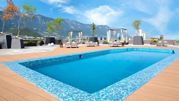 Uma piscina retangular com borda em azulejo, rodeada por um deck de madeira. Há cadeiras de descanso e uma área de estar coberta, com vista para montanhas ao fundo.
