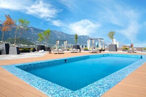 A rectangular swimming pool with a tiled edge, surrounded by wooden decking. There are lounge chairs and a covered seating area with a mountain view in the background. - Camino Real Fashion Drive Monterrey (San Pedro Garza García)