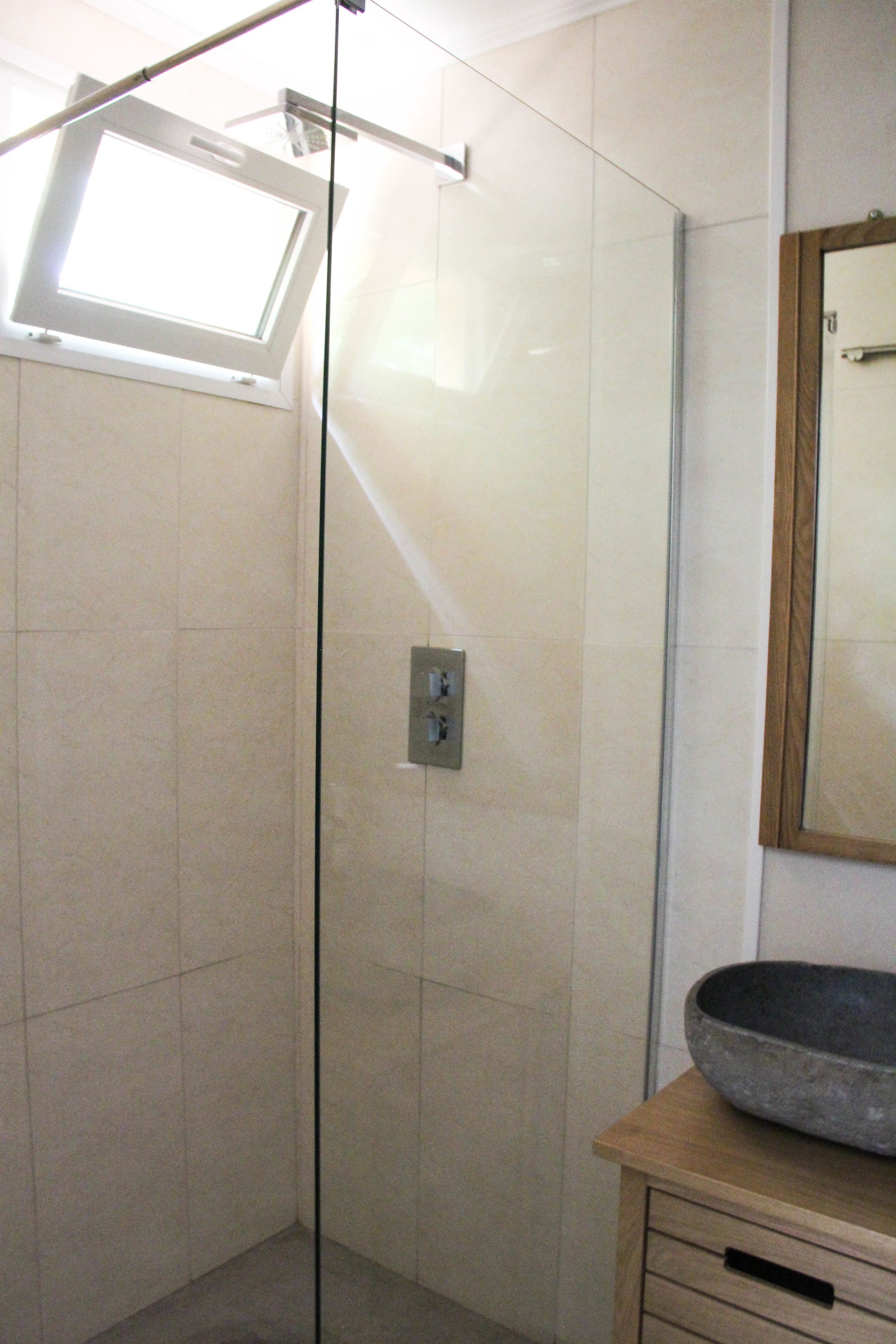 Une salle de bain moderne dotée d’une douche à parois de verre, d’un puits de lumière, d’un meuble-lavabo en bois avec un vasque en pierre et d’un miroir avec un cadre en bois.