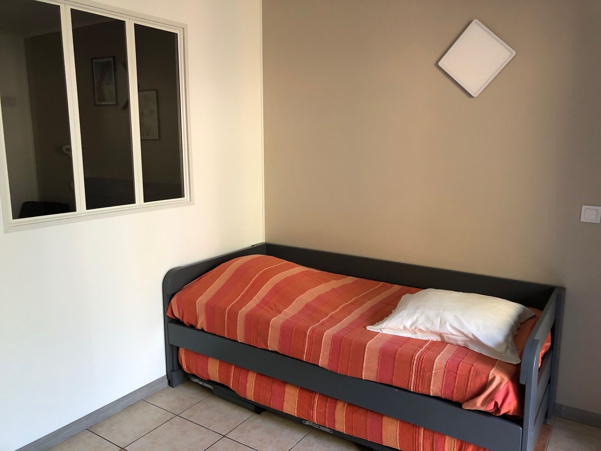 Una cama individual con una colcha a rayas y un almohadón, una ventana con persianas, e interruptor de luz en la pared.