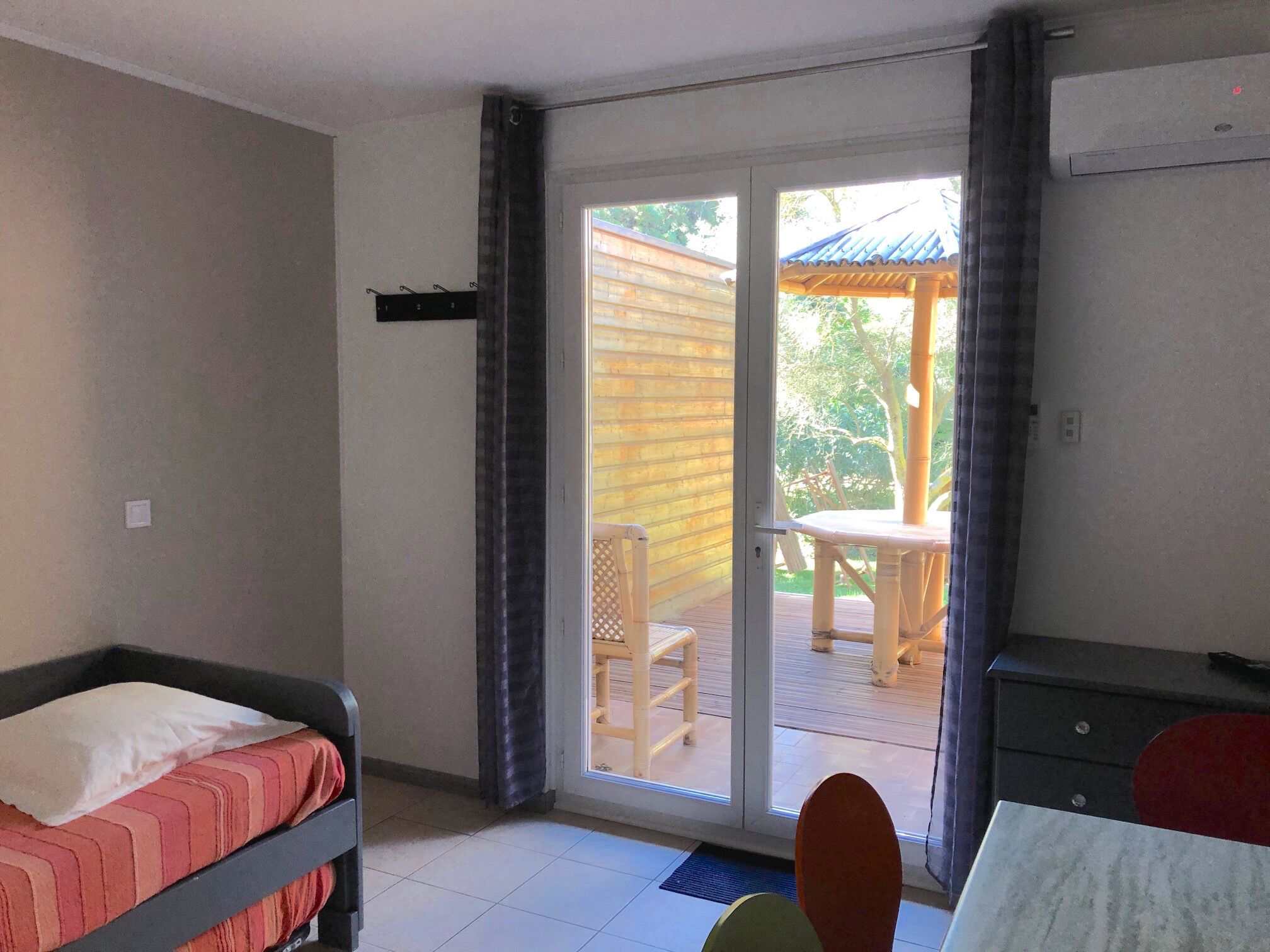 Habitación con cama, escritorio, silla y puerta corrediza de vidrio que da a un espacio exterior con mesa y sillas.