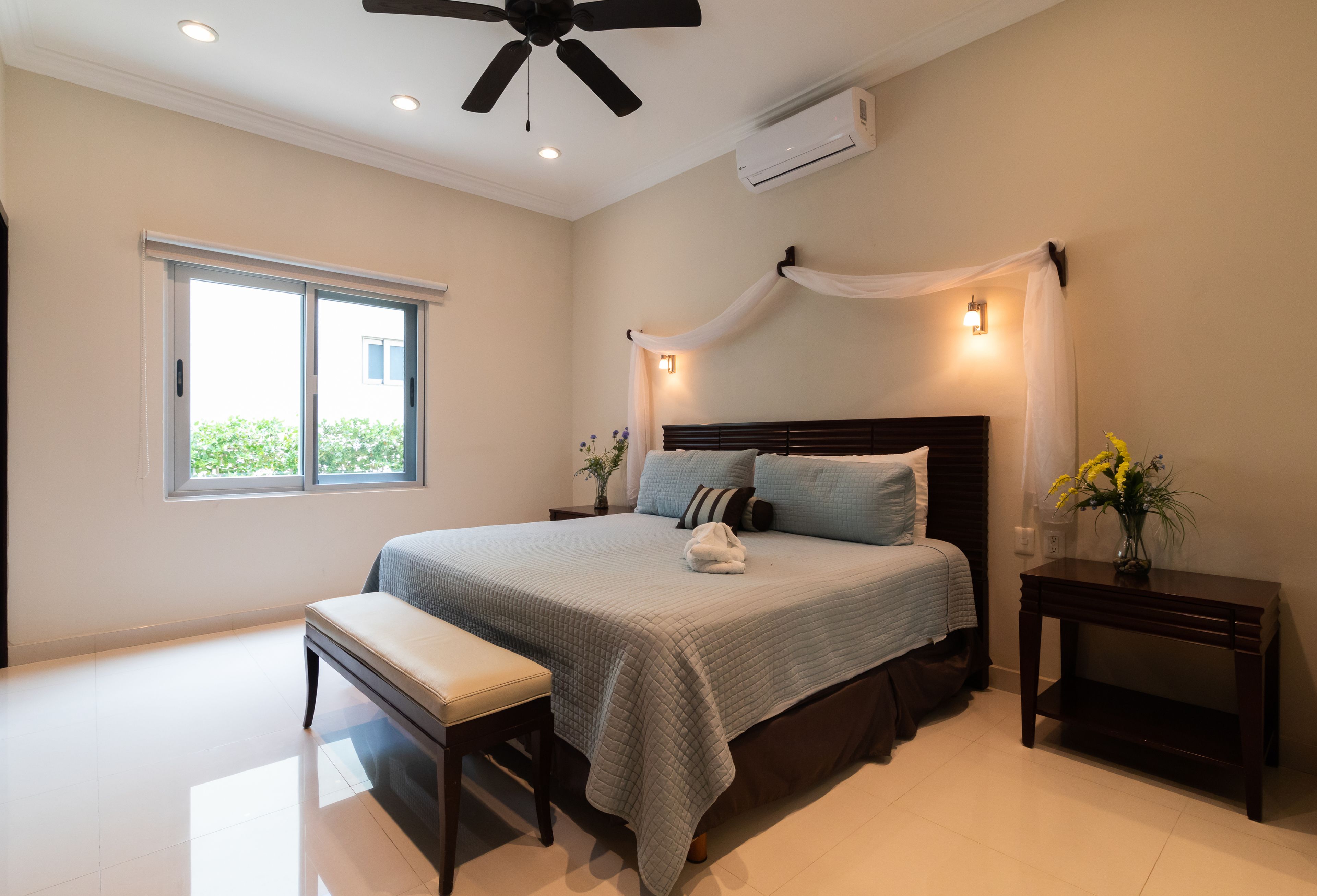 Une chambre à coucher comprenant un lit, un banc, des tables de chevet et un ventilateur de plafond.