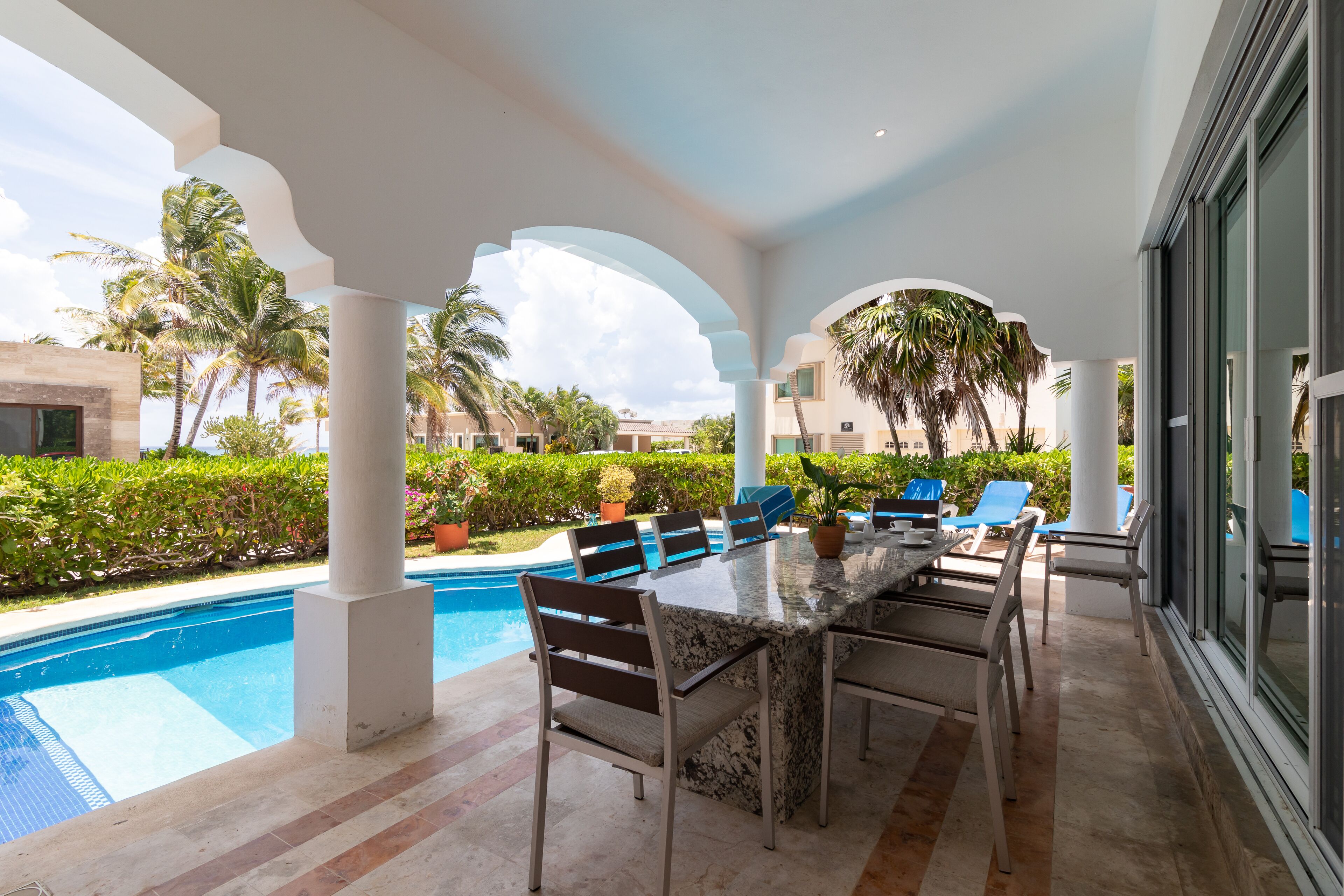 An outdoor pool area with a long dining table and chairs, surrounded by palm trees and a clear sky.